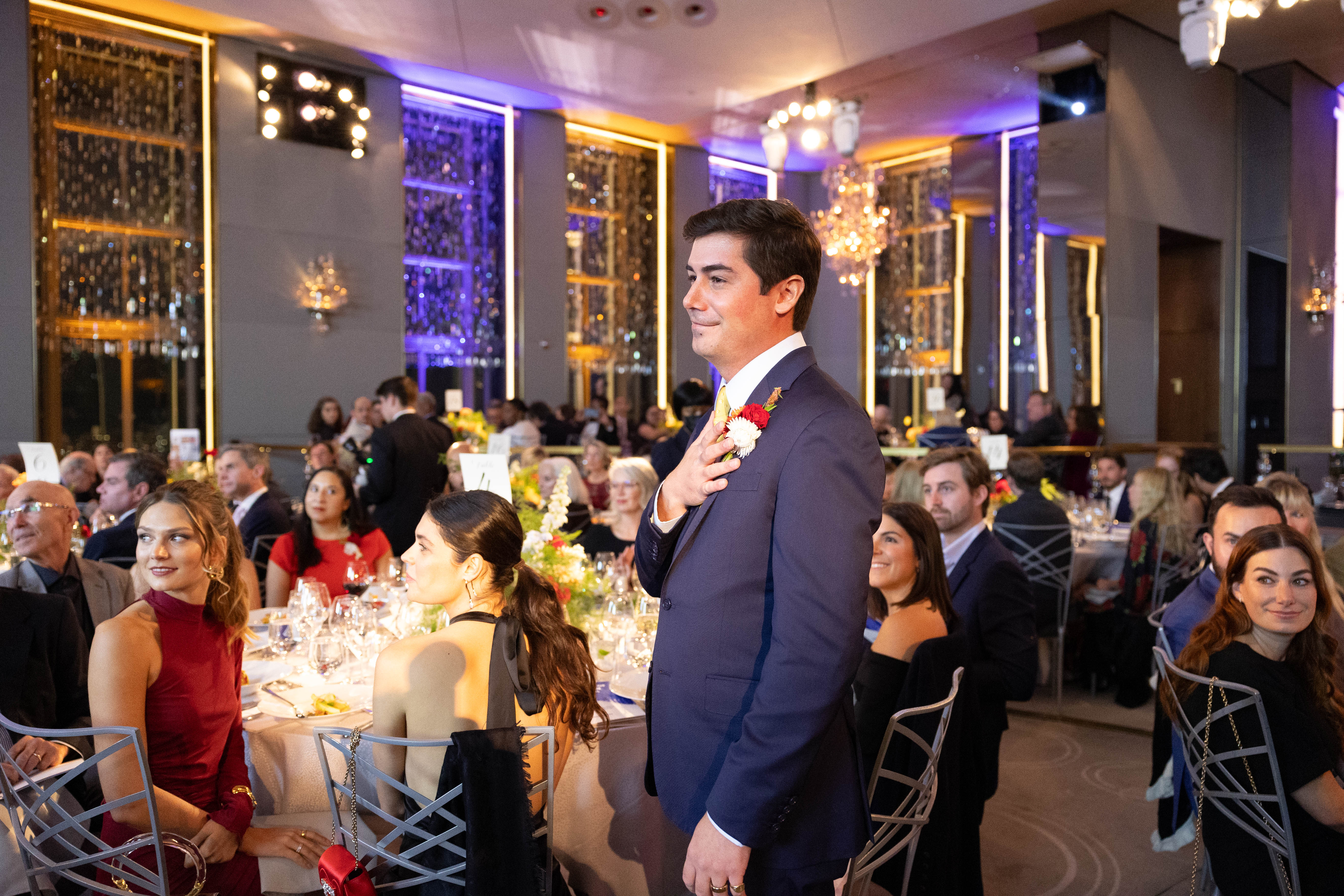 George Merck standing with his hand over his heart after a heartfelt speech  on the floor of the Gala with other guests sitting at their tables and watching. 