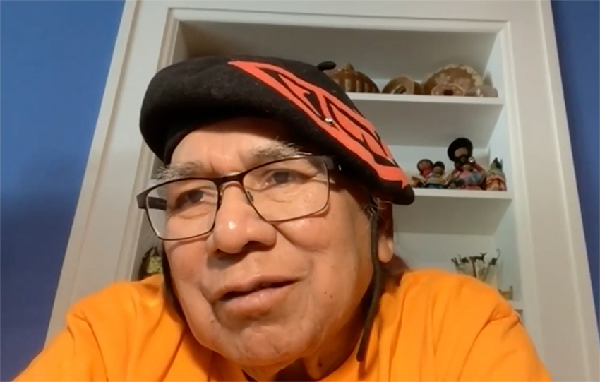 Close-up screenshot of a man wearing glasses, an orange and black beret, and an orange shirt in front of a blue wall with white built-in shelves holding several art objects.