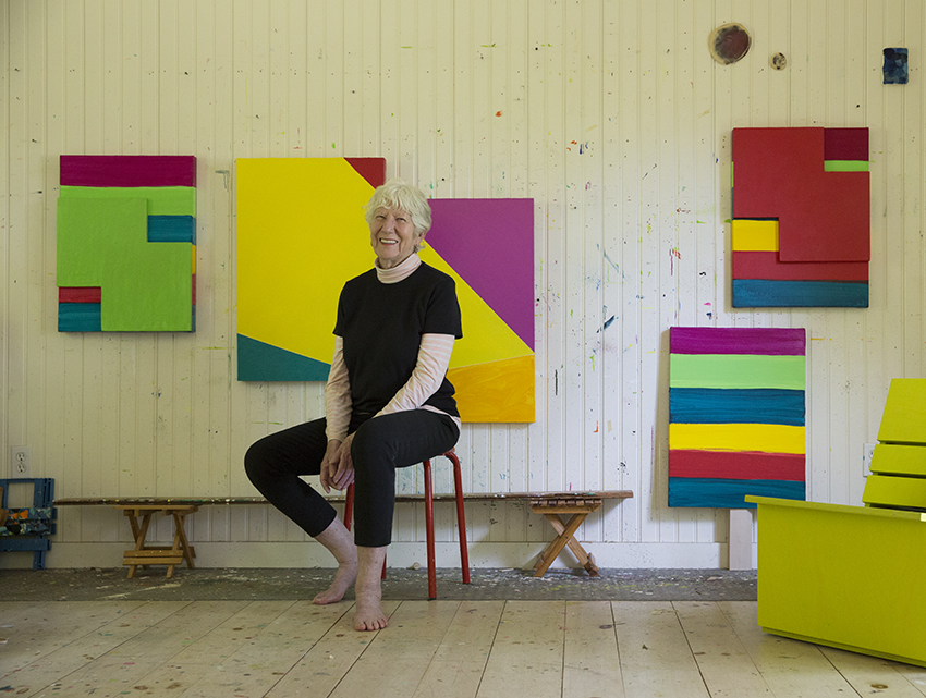 Silver-haired woman dressed in black and barefoot sits on a stool in an art studio surrounded by brightly colored canvases. Photograph by Michael Halsband, courtesy of the Mary Heilmann Studio. 