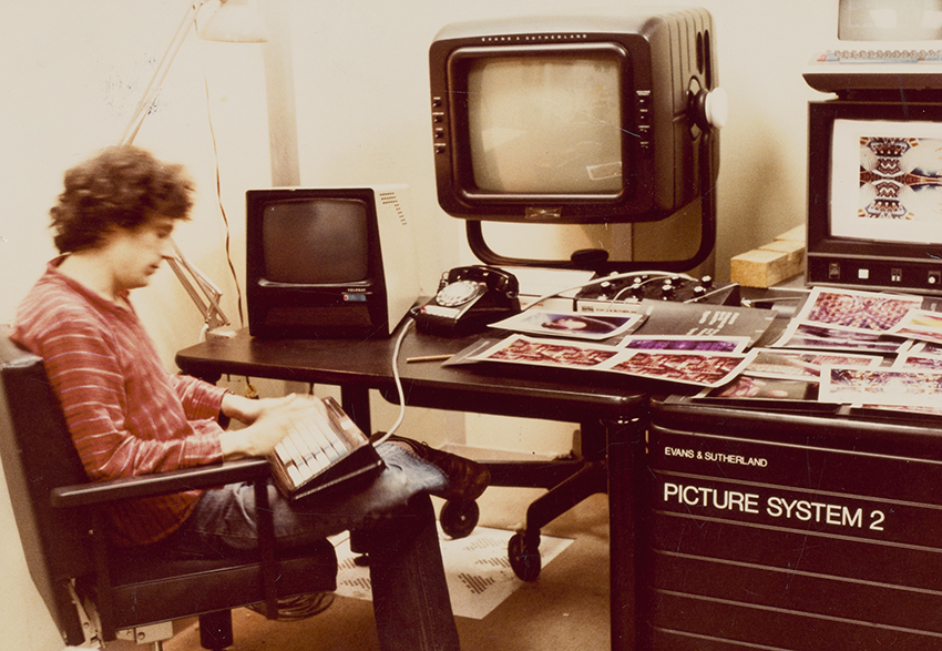 A man in a red striped sweater and jeans sits cross-legged at a desk with early computer models stacked on the desk.