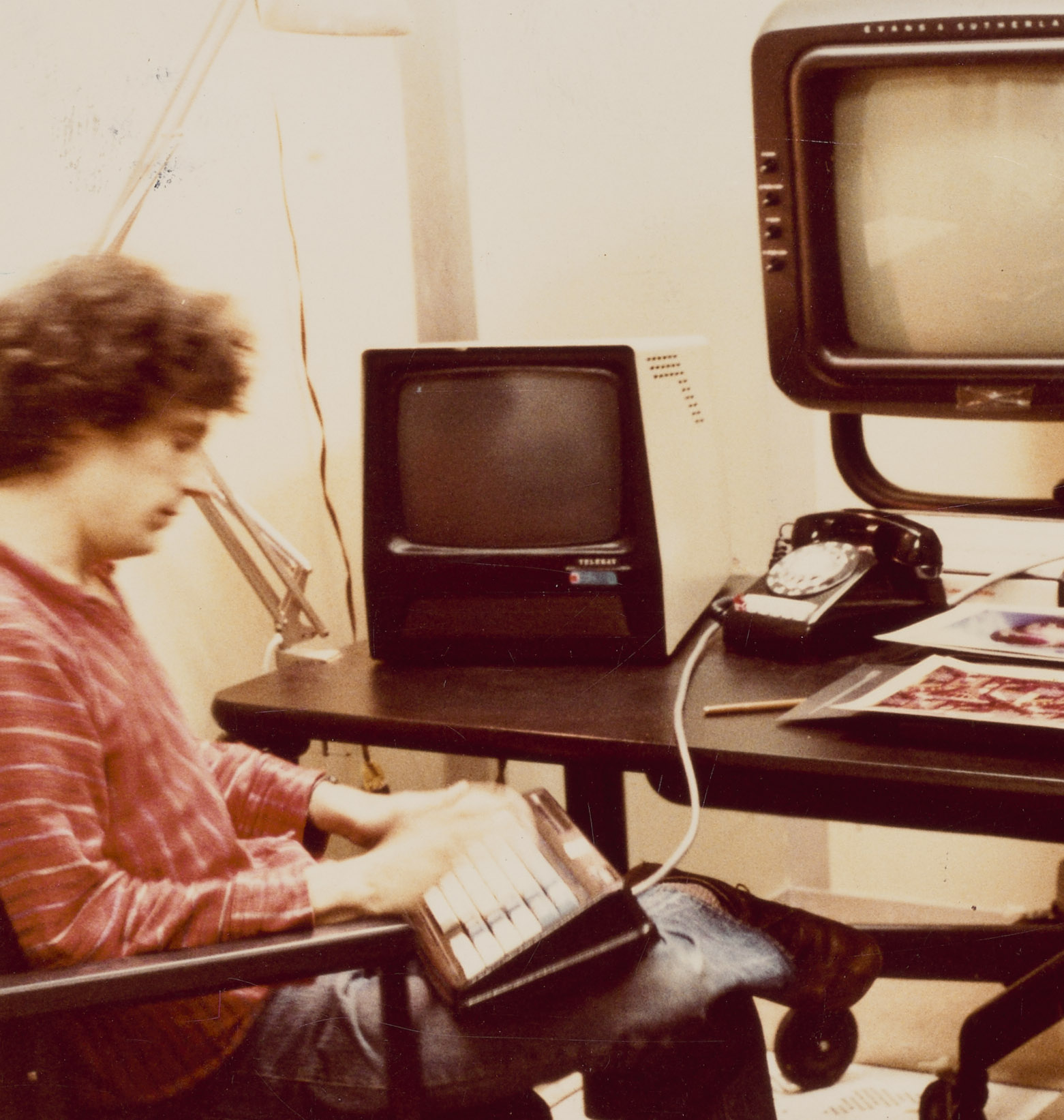 Detail of a color image of a man in a red striped sweater and jeans sitting cross-legged  at a desk with early-model computers stacked on the desk.