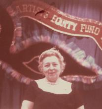 Detail of H. Bella Schaeffer wearing black sitting at a table in front of a dark blue drape with a large-scale black masquerade mask decorated with blue and pink tulle and pink ribbons, and metallic cat eyes.