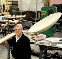 Detail of image where a man stands in a large sculpture studio holding a large spoon, a sculpture component that is roughly 8 to 10 feet long. 