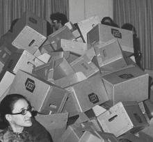 Detail of grayscale image of many people gathered in a room, surrounding empty boxes stacked haphazardly. 